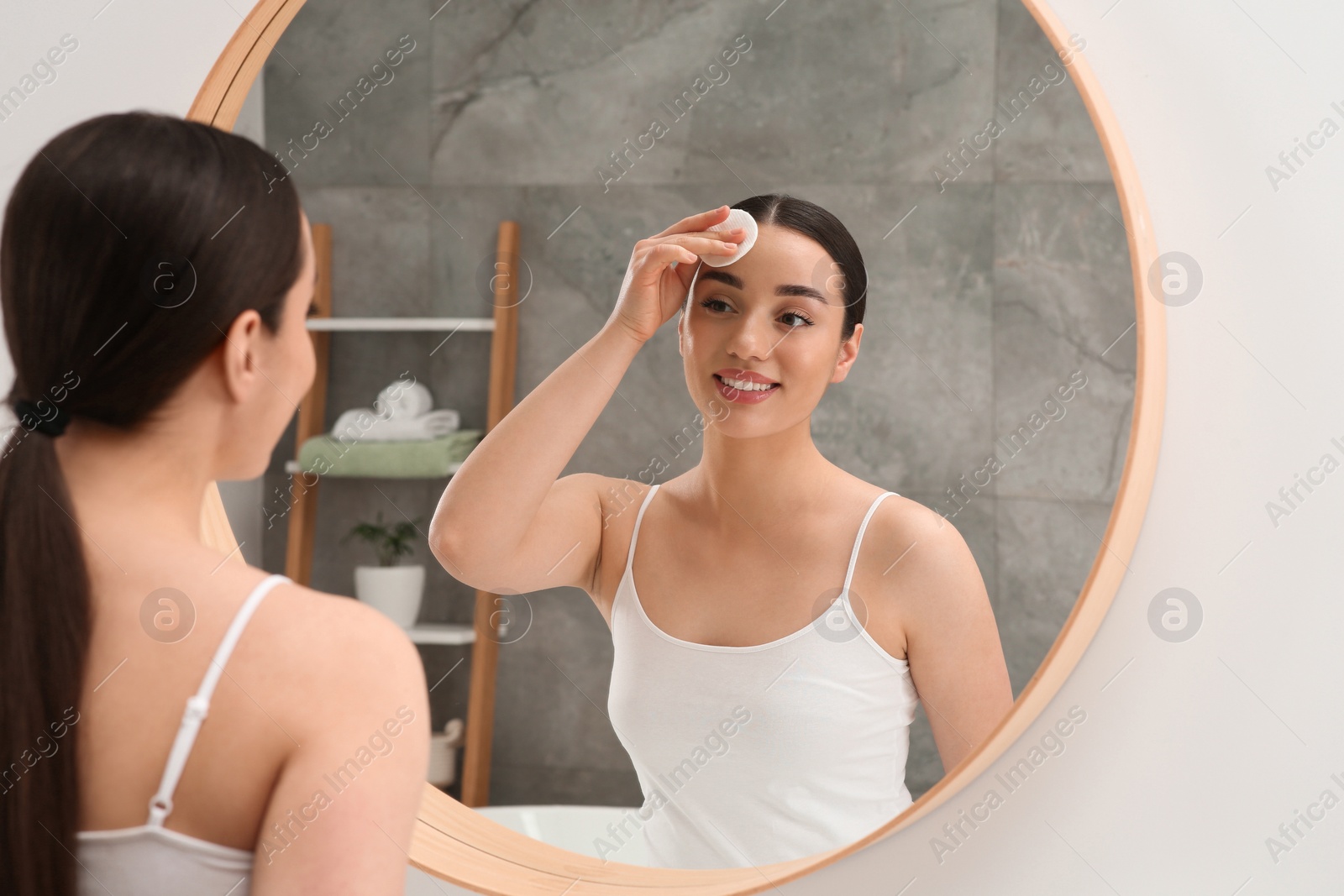 Photo of Beautiful woman removing makeup with cotton pad near mirror indoors