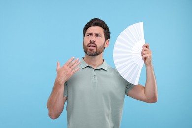 Unhappy man with hand fan suffering from heat on light blue background
