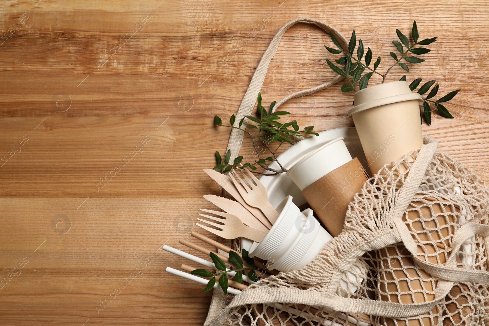 Photo of Net bag with disposable tableware and green twigs on wooden background, top view. Space for text