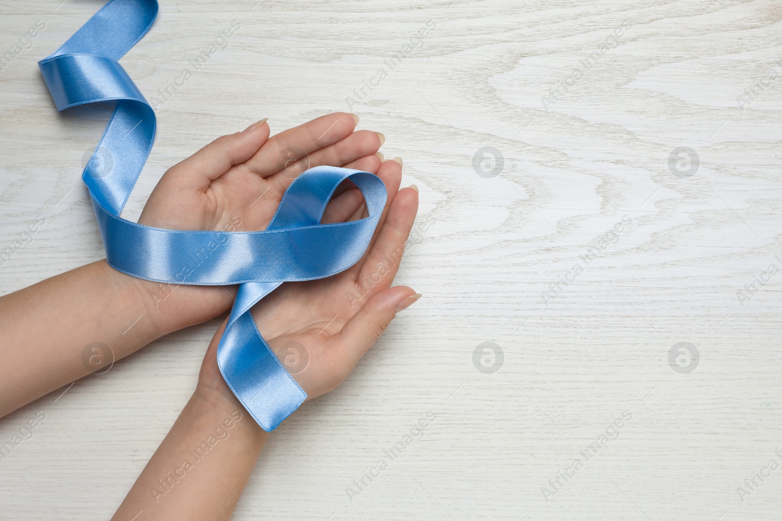 Photo of Woman holding light blue awareness ribbon at white wooden table, top view. Space for text