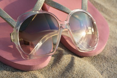 Photo of Stylish sunglasses and pink flip flops on sandy beach, closeup