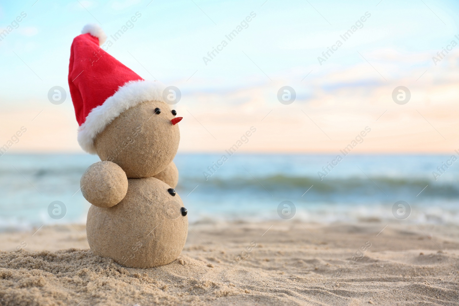 Photo of Snowman made of sand with Santa hat on beach near sea, space for text. Christmas vacation