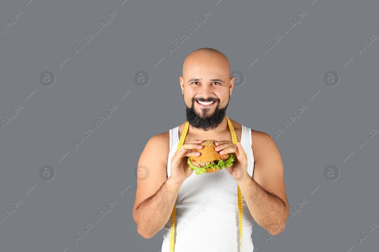 Photo of Overweight man with hamburger and measuring tape on gray background