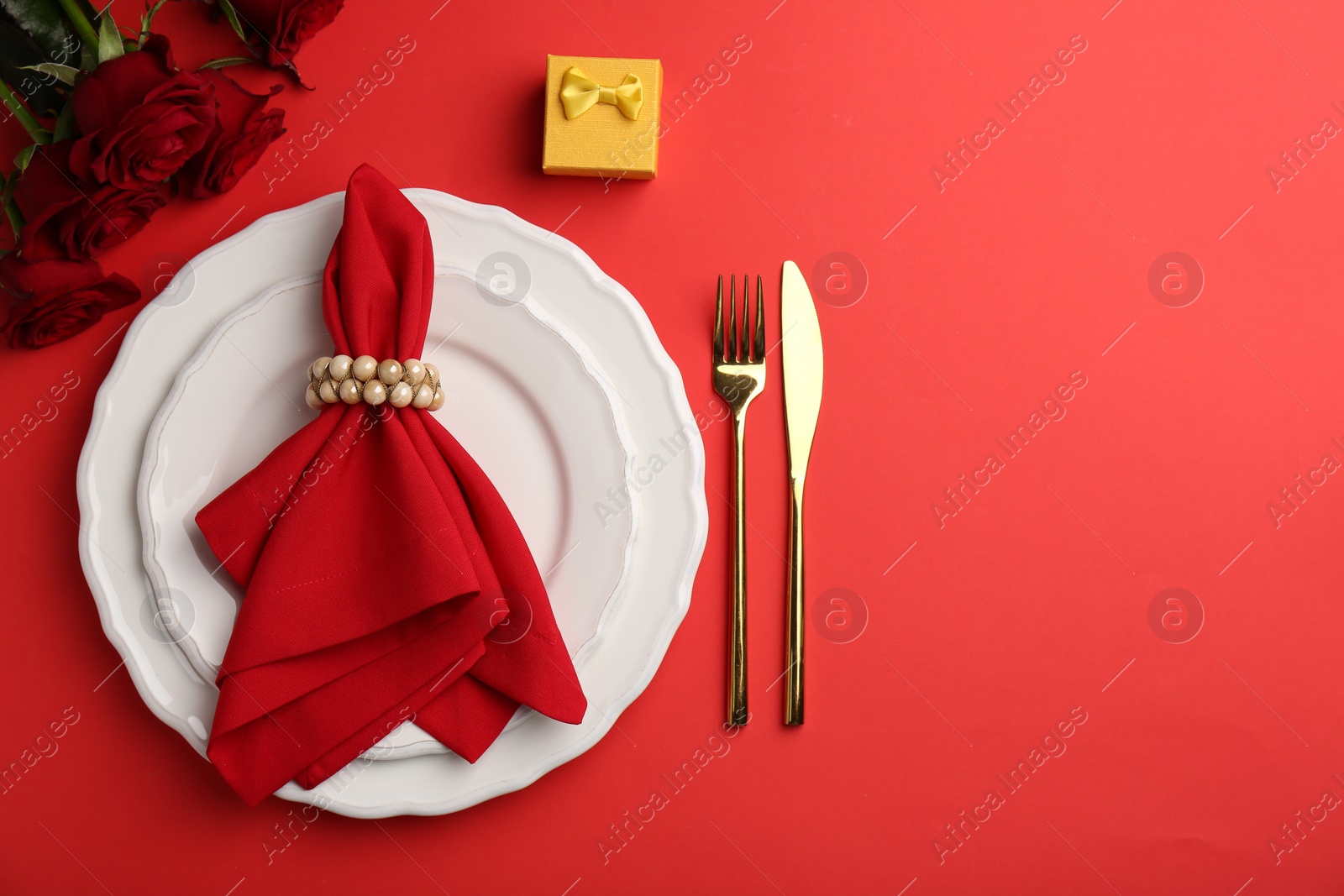Photo of Place setting with gift box and bouquet of roses for romantic dinner on red table, flat lay. Space for text