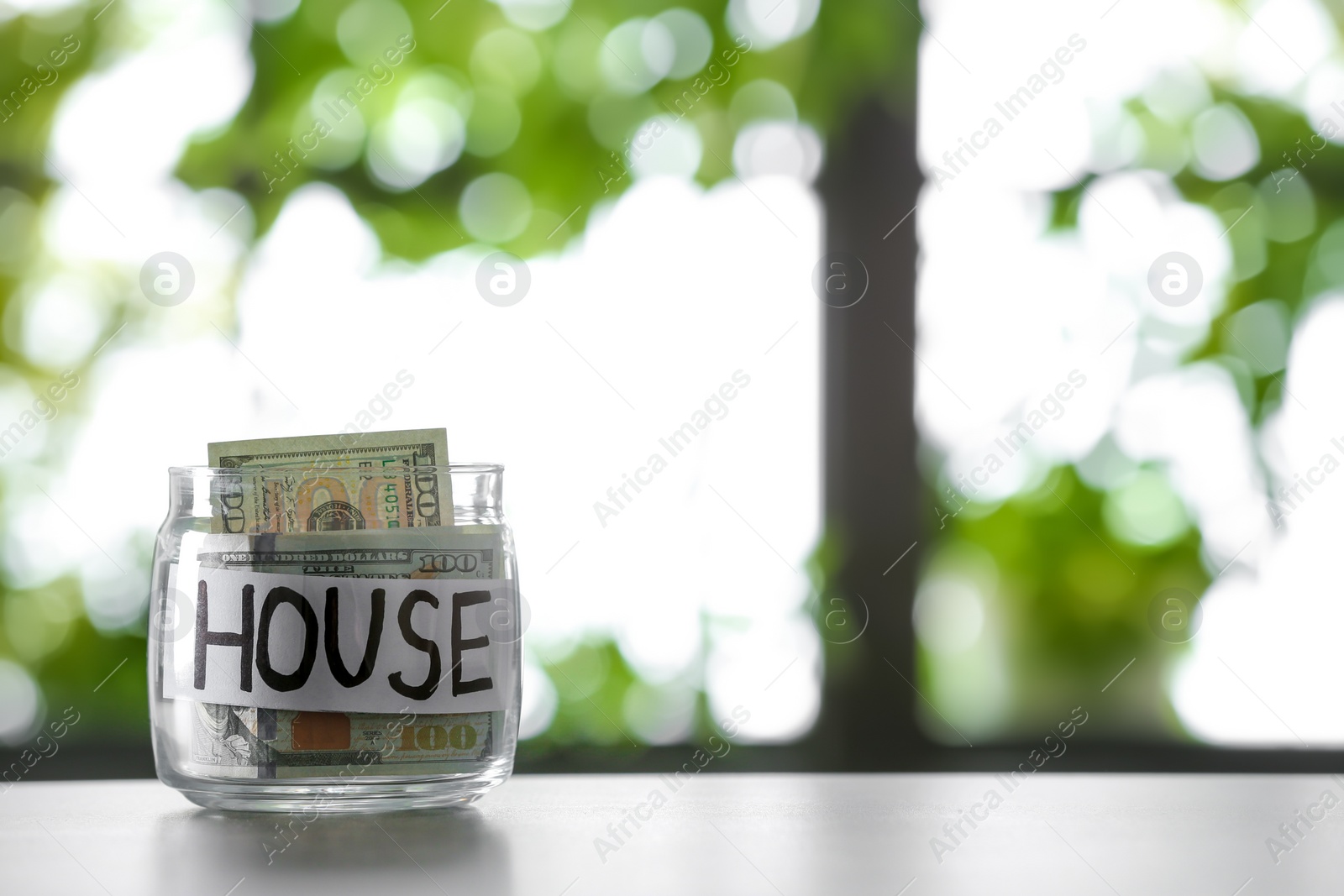 Photo of Glass jar with money and word HOUSE on table against blurred background, space for text