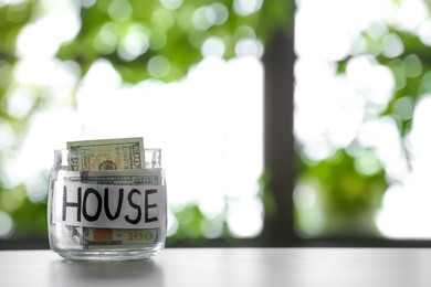 Glass jar with money and word HOUSE on table against blurred background, space for text