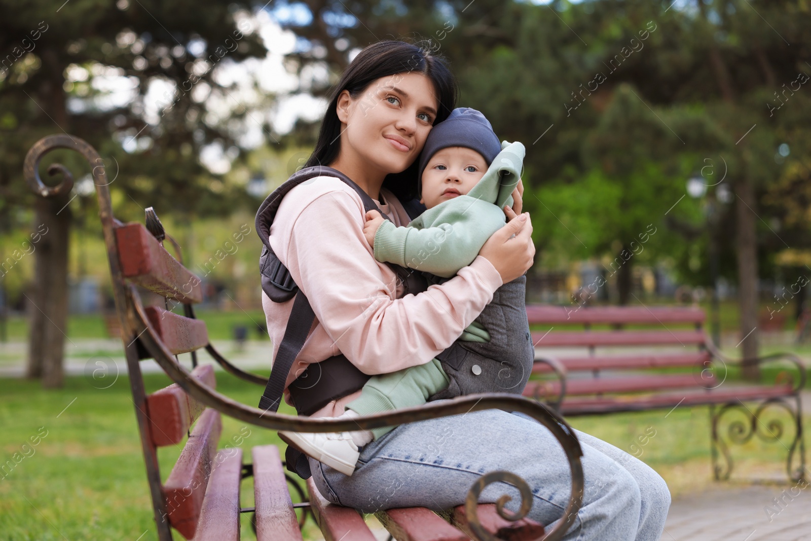 Photo of Mother holding her child in sling (baby carrier) on bench in park