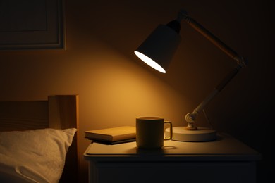 Photo of Stylish modern desk lamp, books and cup of drink on white nightstand in dark bedroom
