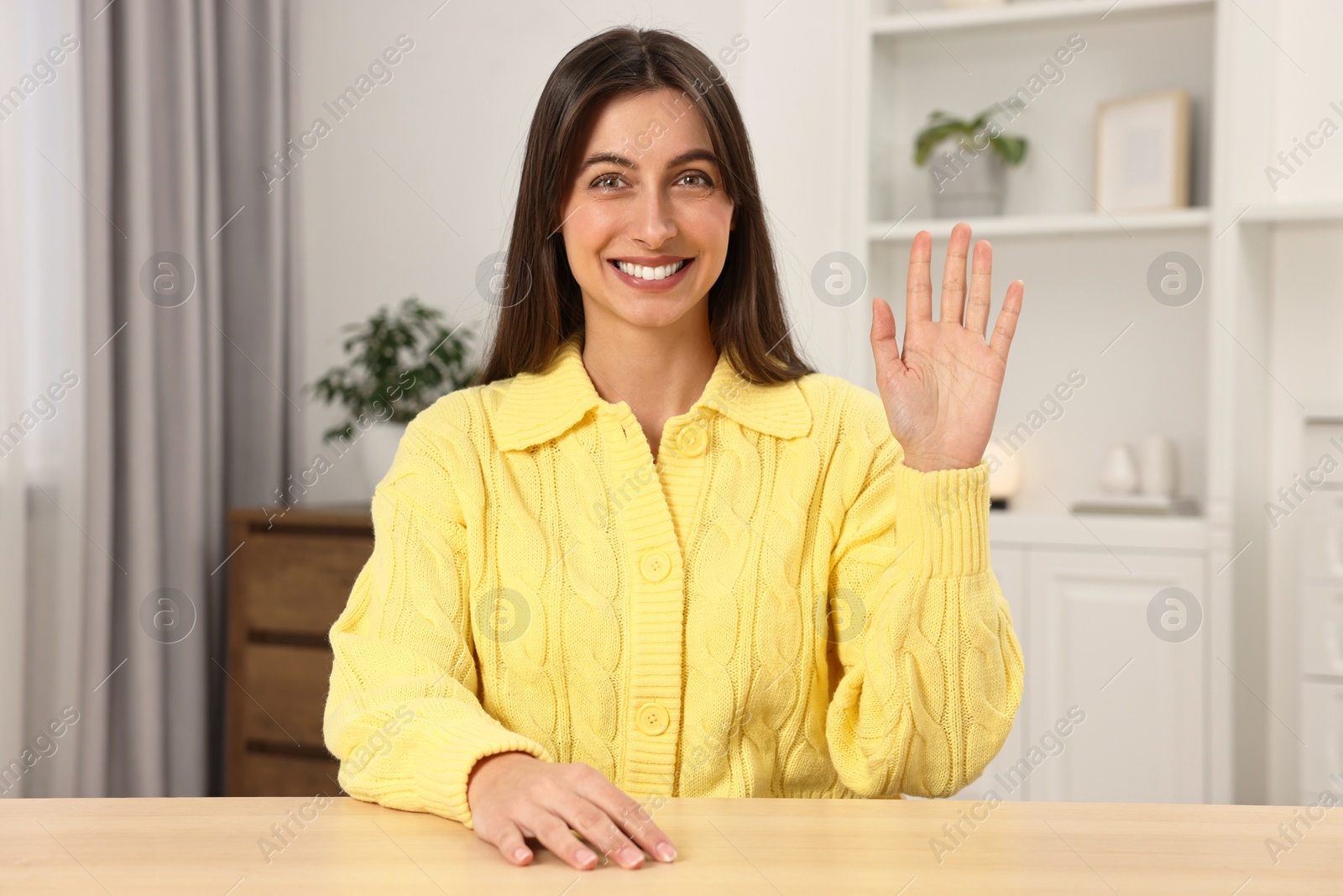 Photo of Happy woman waving hello at table indoors
