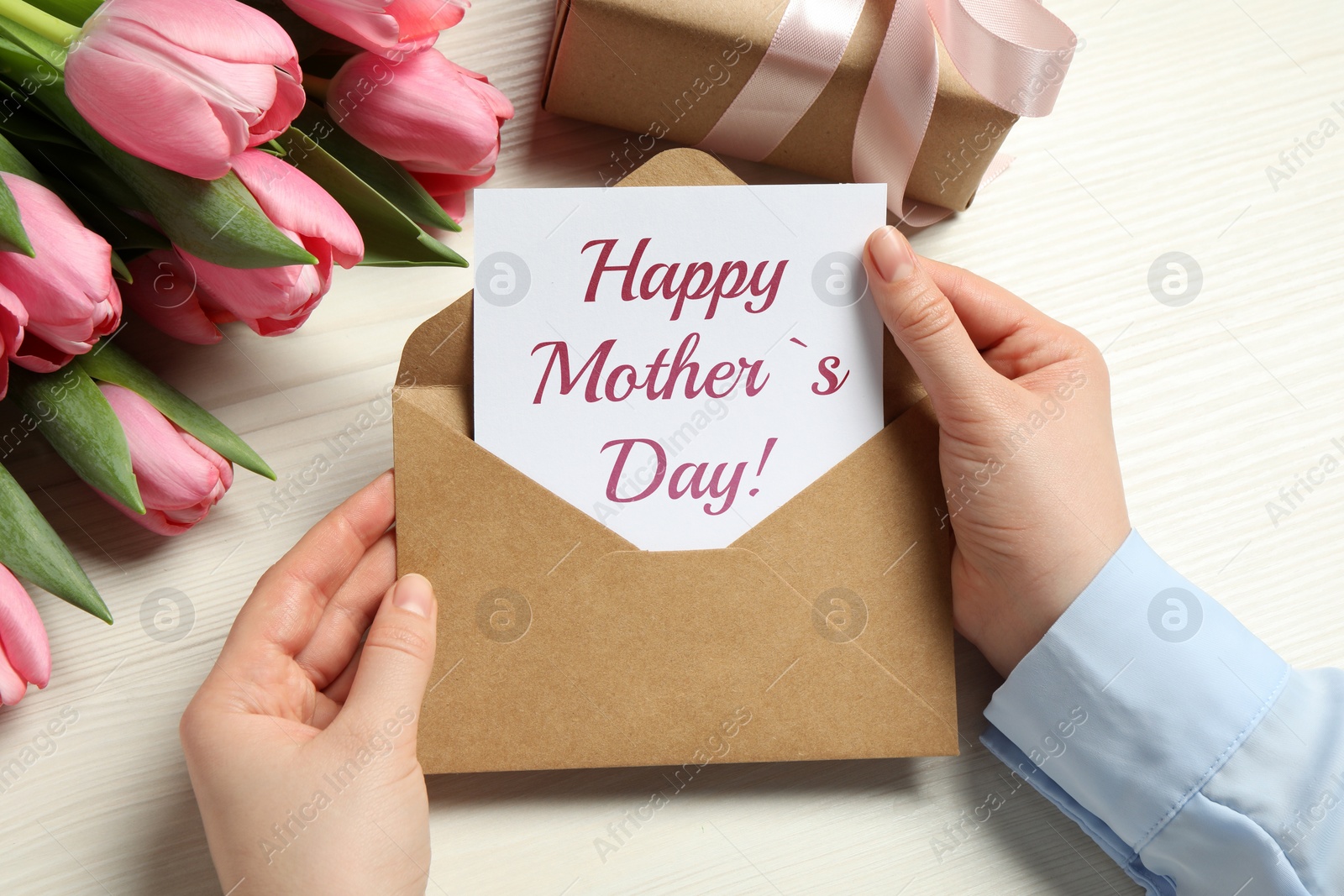 Image of Happy Mother's Day. Woman taking greeting card out of envelope, closeup. Gift box and bouquet of beautiful flowers on white wooden table
