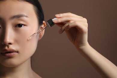 Beautiful young woman applying cosmetic serum onto her face on brown background, closeup