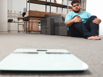 Photo of Depressed overweight man looking at scales in living room