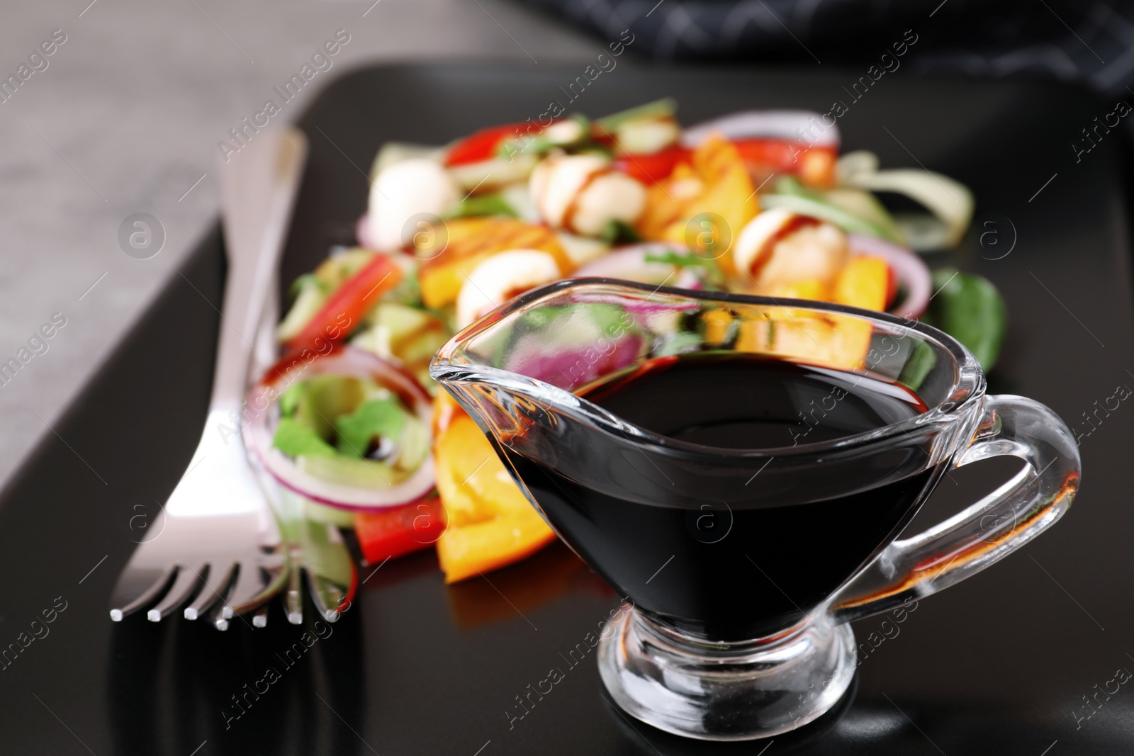 Photo of Gravy boat of balsamic vinegar on plate with vegetable salad, closeup