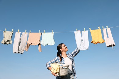Photo of Smiling woman hanging baby clothes with clothespins on washing line for drying against blue sky outdoors