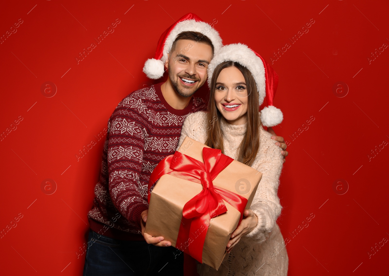 Photo of Couple in Christmas sweaters and Santa hats with gift box on red background