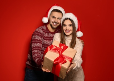 Couple in Christmas sweaters and Santa hats with gift box on red background
