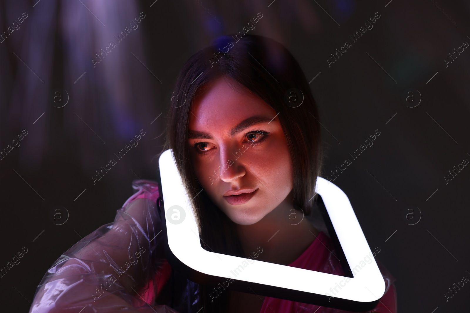 Photo of Fashionable portrait of beautiful woman wearing transparent coat with square lamp on dark background in neon lights