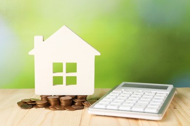 Photo of House model, calculator and coins on wooden table against blurred green background, space for text