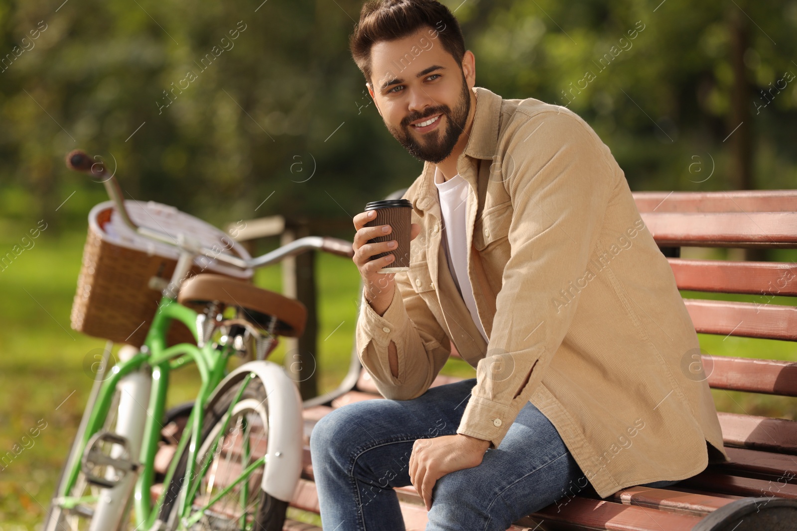 Photo of Young man sitting on bench and holding cup of coffee in park, space for text