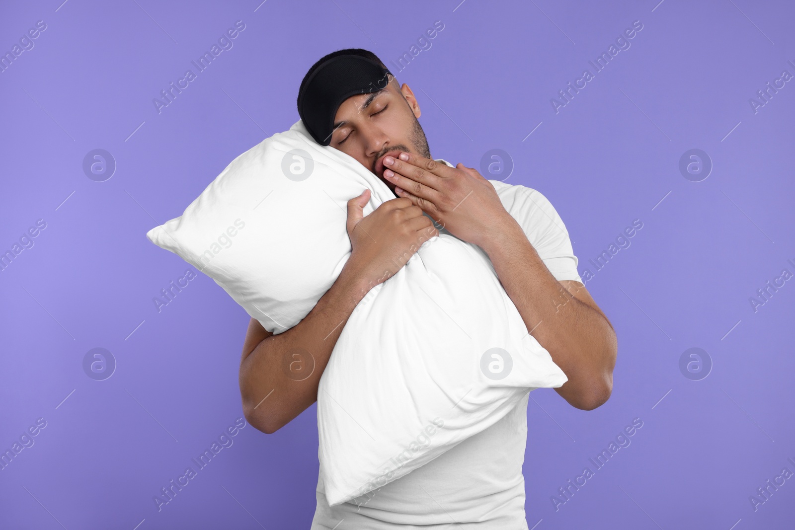 Photo of Tired man with pillow and sleep mask yawning on violet background. Insomnia problem