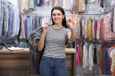 Photo of Dry-cleaning service. Happy woman holding hanger with jacket in plastic bag and showing ok gesture indoors