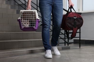 Photo of Travel with pet. Man holding carrier with cute cat and bag indoors, closeup