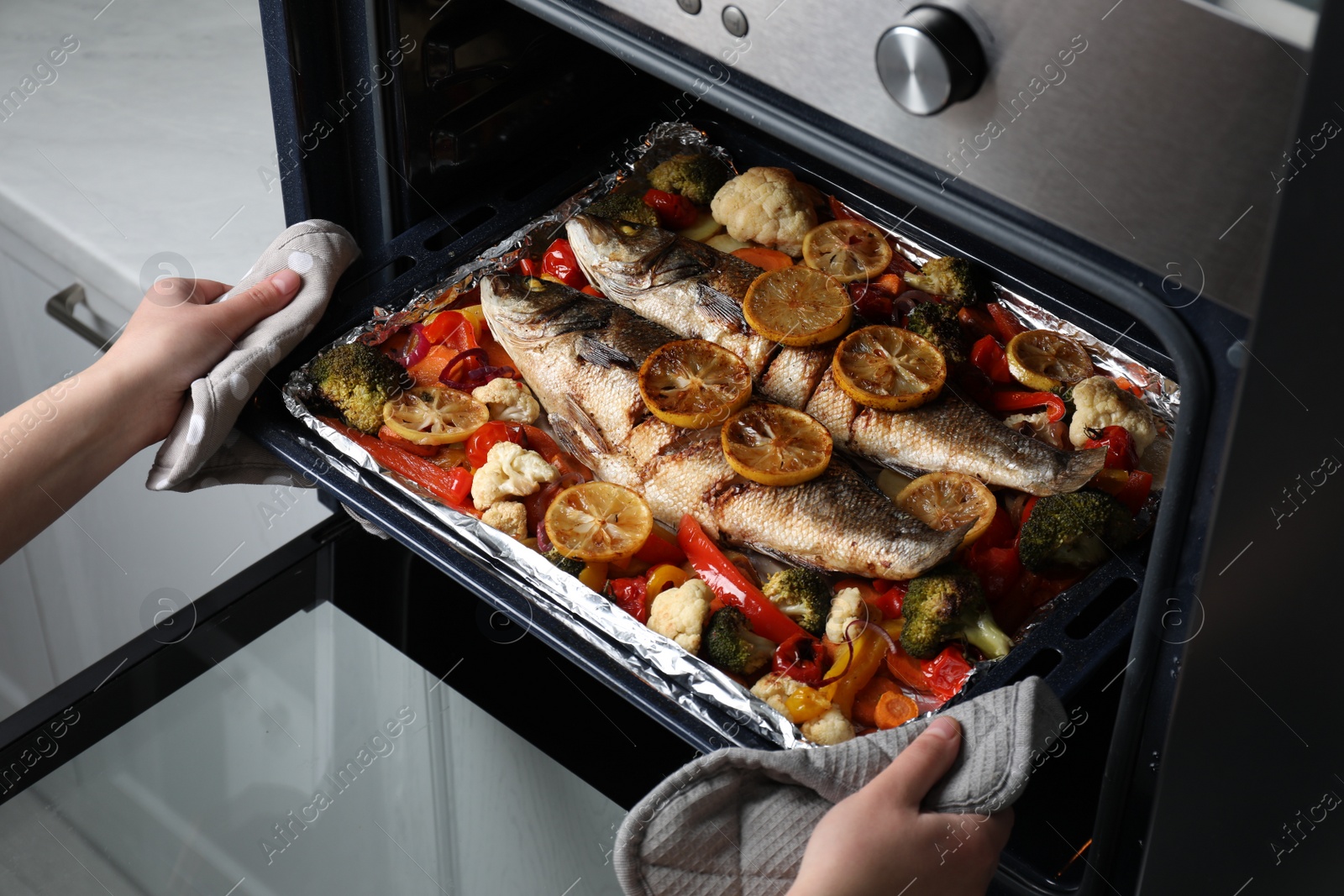 Photo of Woman taking out baking tray with sea bass fish and vegetables from oven, closeup