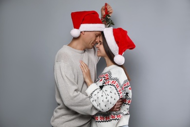 Happy couple in Santa hats standing under mistletoe bunch on grey background