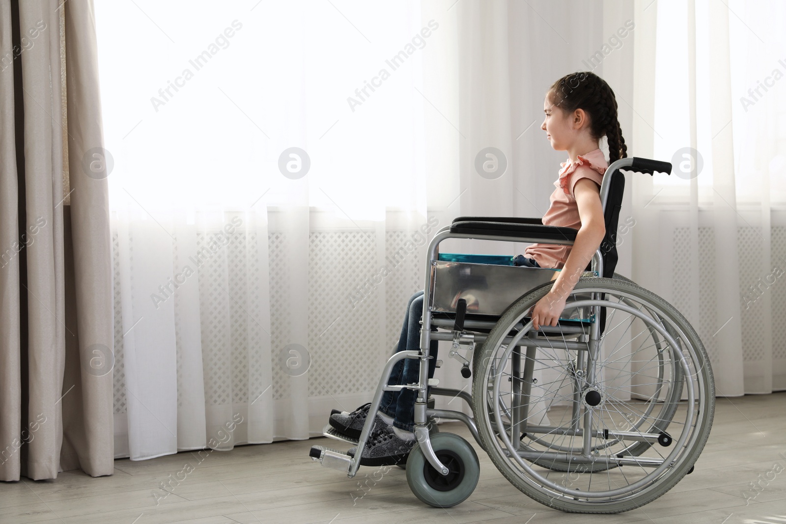 Photo of Little girl in wheelchair near window indoors. Space for text