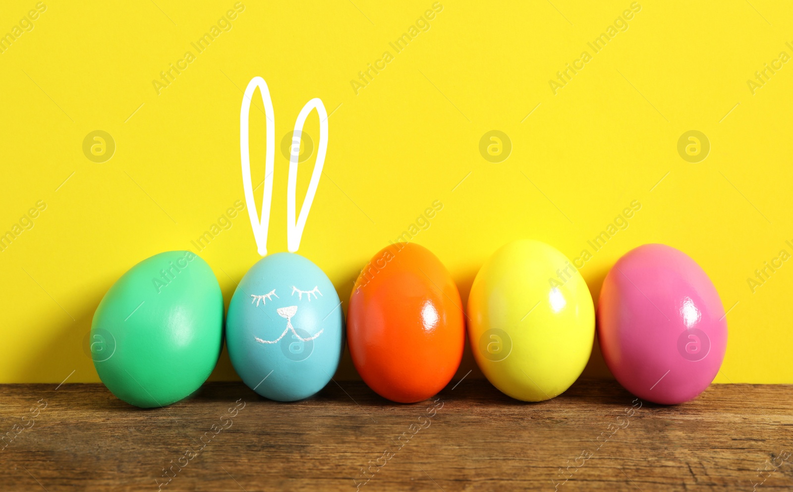 Image of Blue egg with drawn face and ears as Easter bunny among others on wooden table against yellow background