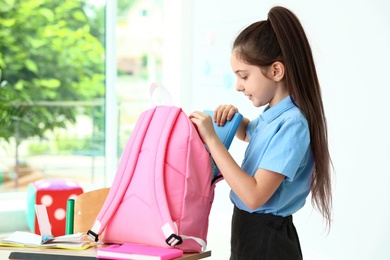 Photo of Little girl putting school stationery into backpack at desk in classroom