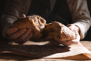 Photo of Man breaking loaf of fresh bread at wooden table, closeup