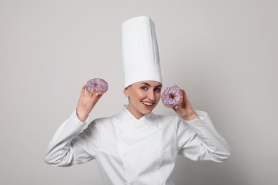 Photo of Happy professional confectioner in uniform holding delicious doughnuts on light grey background
