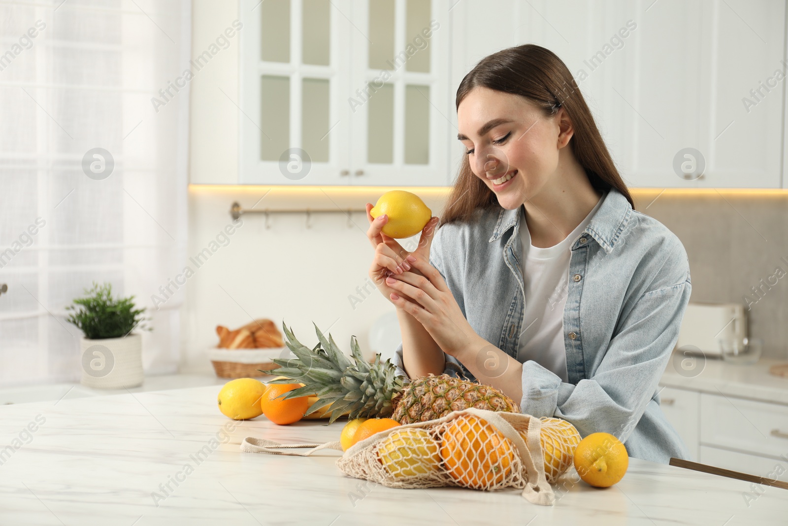 Photo of Woman with lemon and string bag of fresh fruits at light marble table in kitchen
