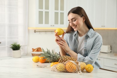 Woman with lemon and string bag of fresh fruits at light marble table in kitchen