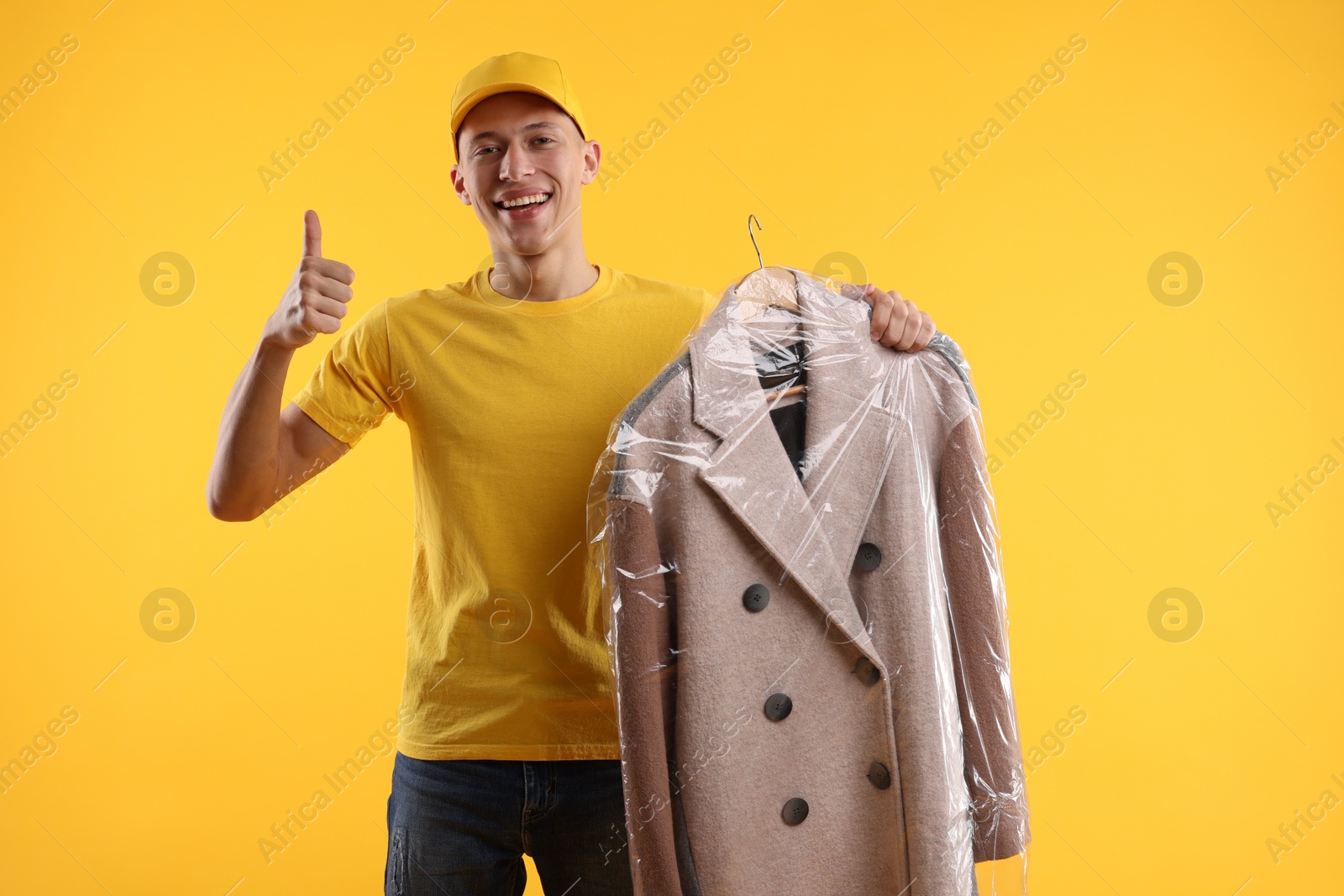 Photo of Dry-cleaning delivery. Happy courier holding coat in plastic bag and showing thumbs up on orange background