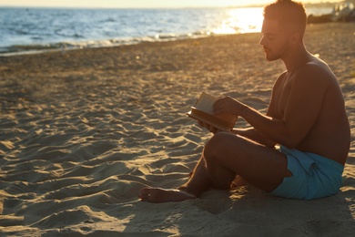 Young man reading book on sandy beach near sea