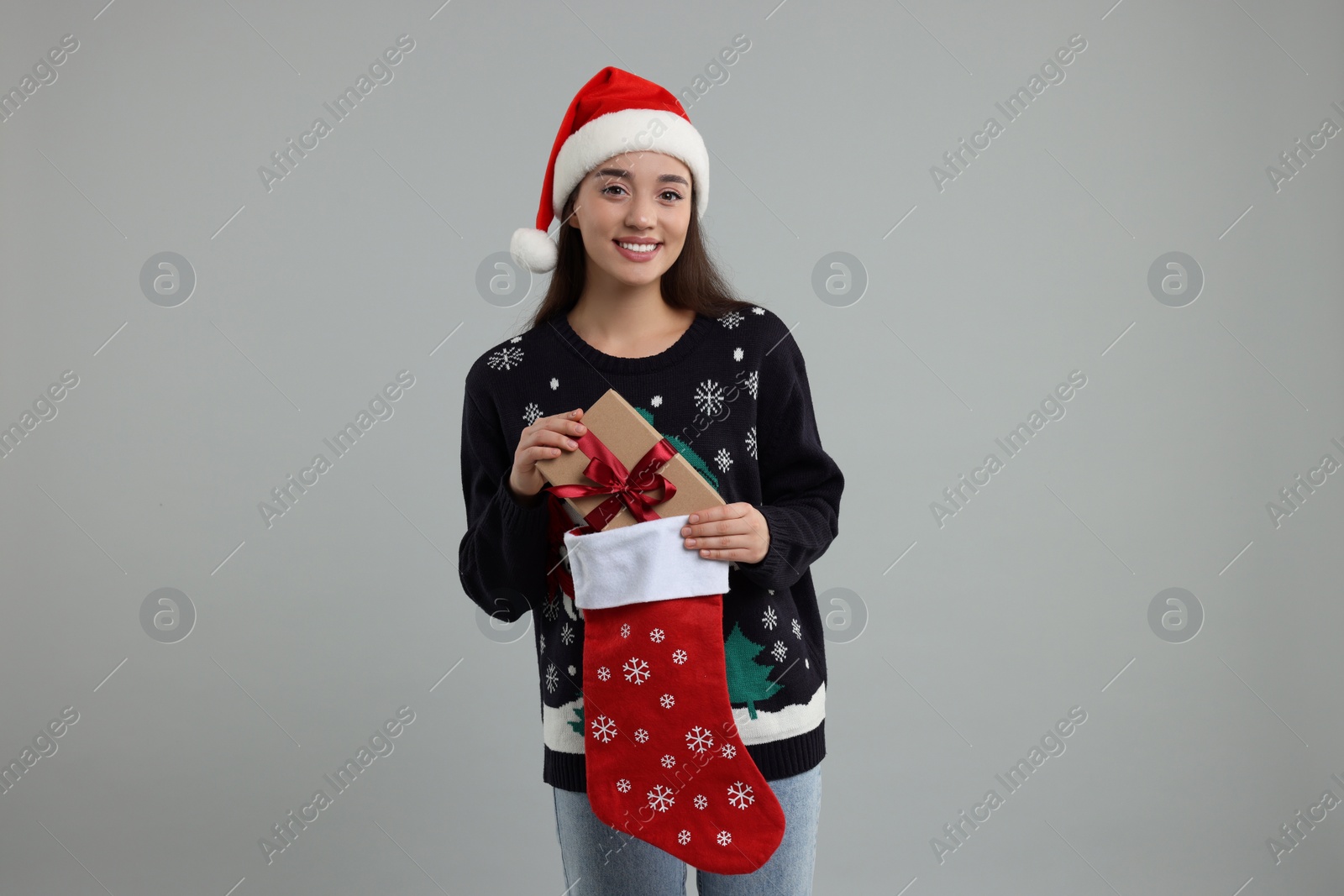 Photo of Young woman in Christmas sweater and Santa hat taking gift from stocking on grey background