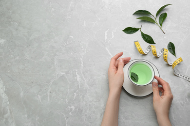 Photo of Woman with cup of herbal diet tea and measuring tape at light table, top view. Space for text