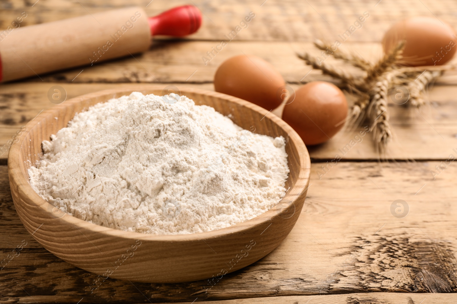 Photo of Bowl with wheat flour near eggs and spikelets on wooden table. Space for text