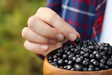 Woman putting bilberry into wooden bowl on blurred background, closeup