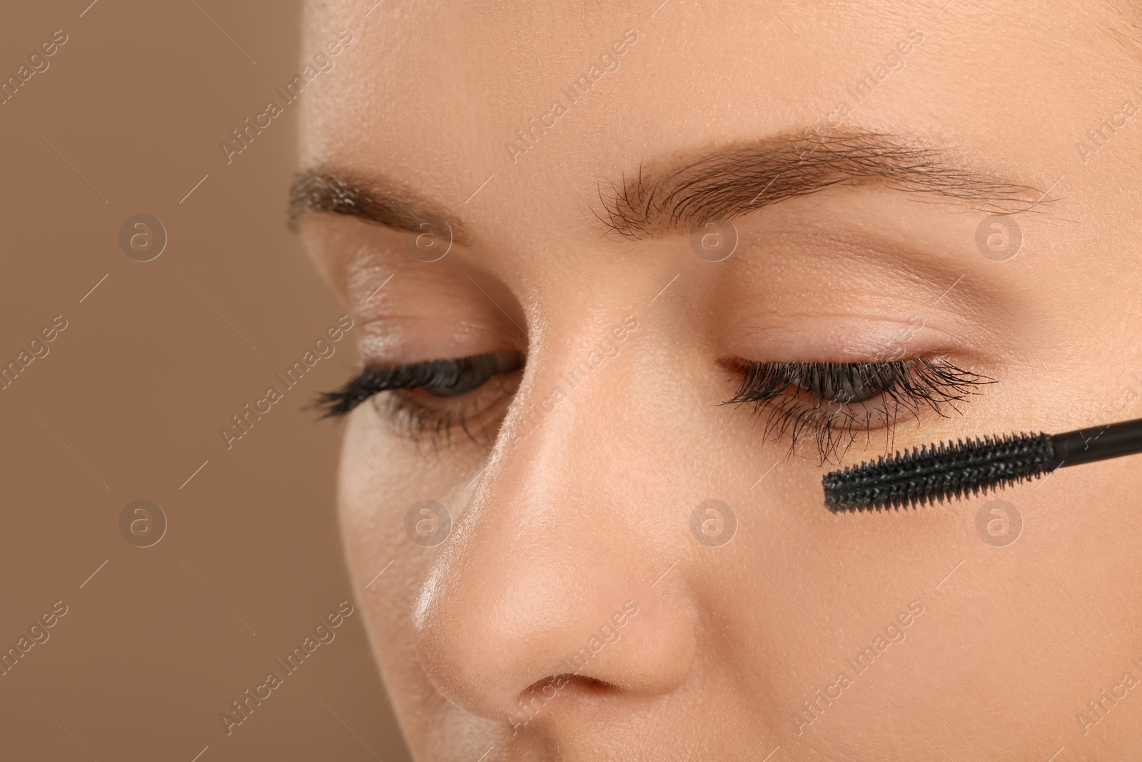 Photo of Woman applying mascara onto eyelashes against light brown background, closeup