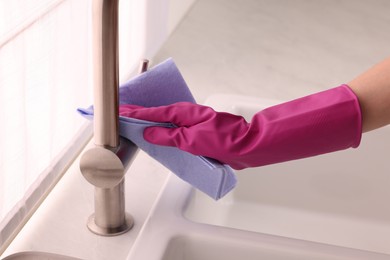 Photo of Woman in gloves cleaning faucet of kitchen sink with rag, closeup