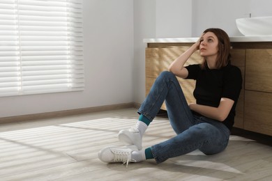 Photo of Sad young woman sitting on floor in bathroom, space for text