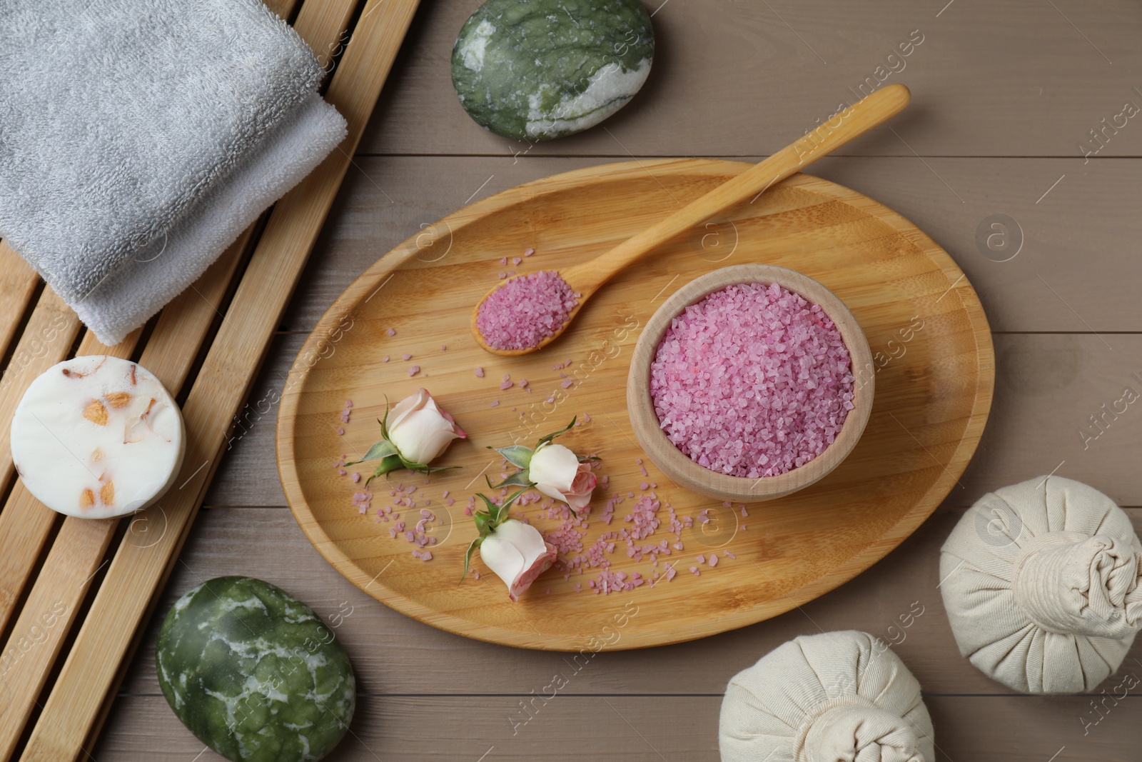 Photo of Bowl of pink sea salt, roses, spa stones, herbal massage bags and towels on wooden table, flat lay