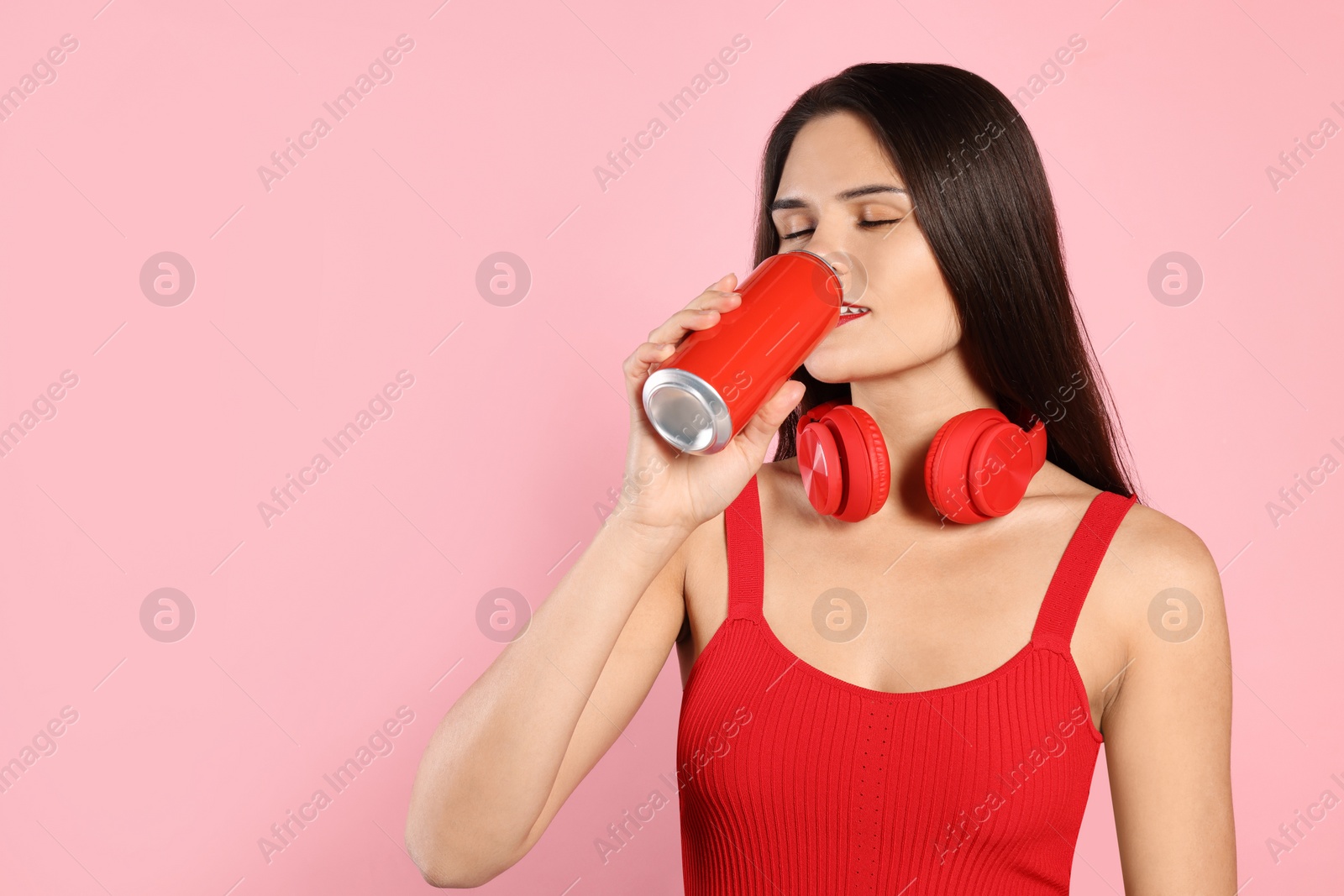 Photo of Beautiful young woman with headphones drinking from tin can on pink background. Space for text