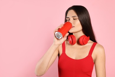 Beautiful young woman with headphones drinking from tin can on pink background. Space for text