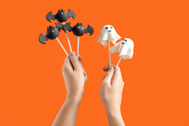 Photo of Woman with delicious ghost and bat shaped cake pops on orange background, closeup. Halloween celebration