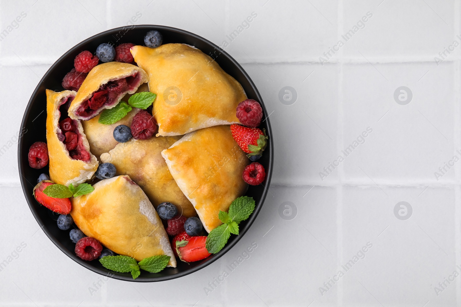 Photo of Bowl with delicious samosas, berries and mint leaves on white tiled table, top view. Space for text