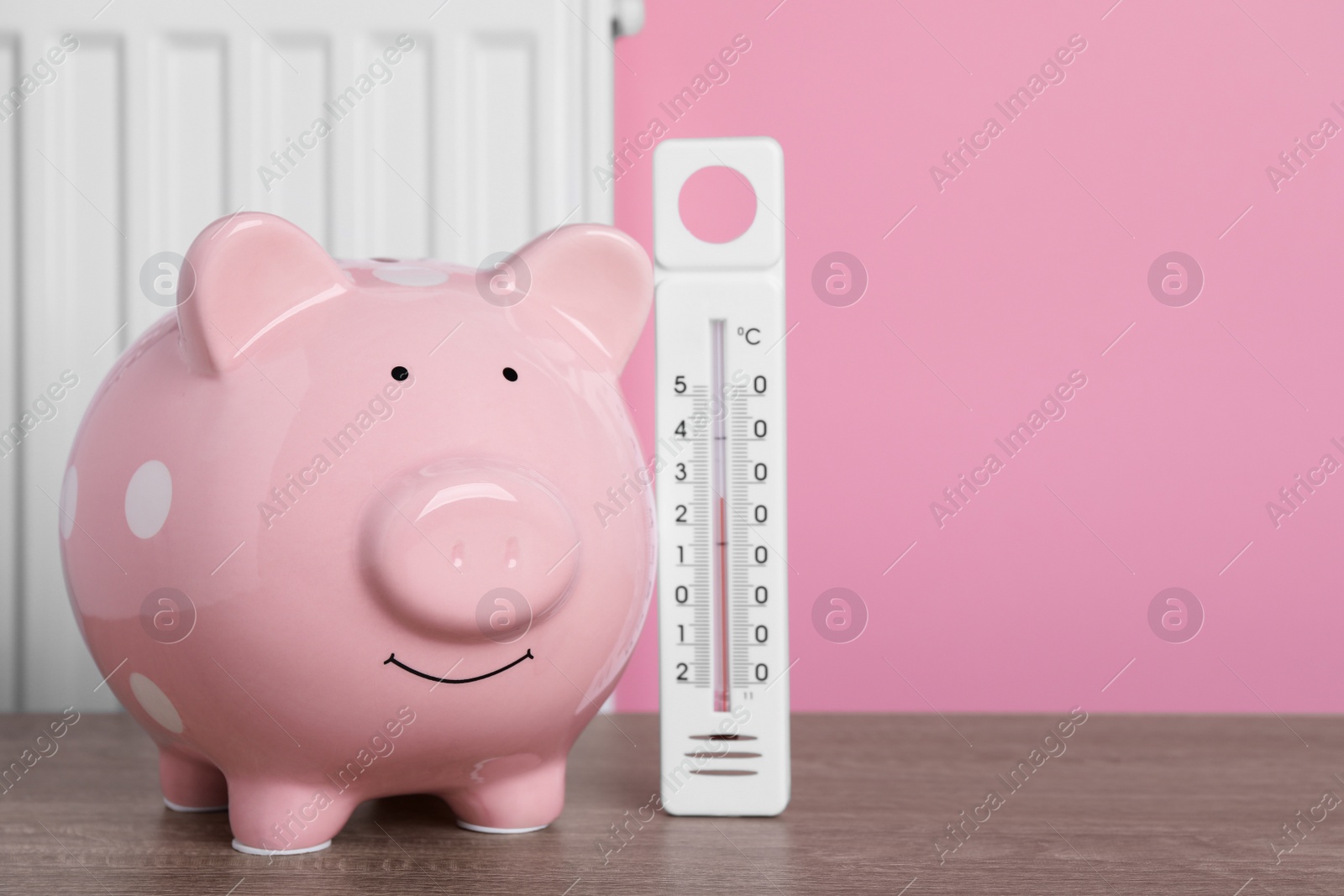 Photo of Piggy bank with thermometer on wooden table near heating radiator. Space for text